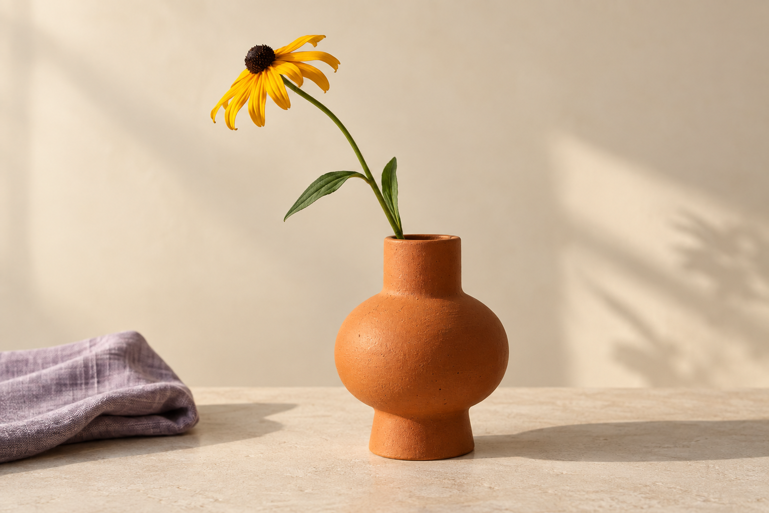A terracotta vase with one yellow flower on a pale surface, with a folded purple cloth at lower left and a plant shadow on the wall at right.